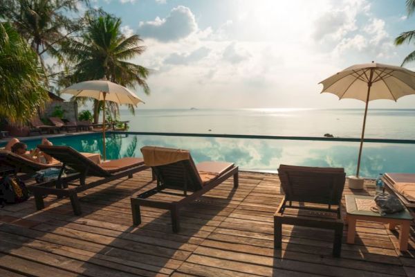 A tropical poolside scene with lounge chairs, umbrellas, and a serene view of the ocean under a partly cloudy sky.