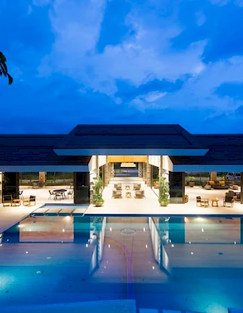 A luxurious poolside view at night, featuring a modern building with outdoor seating and illuminated surroundings under a deep blue sky.
