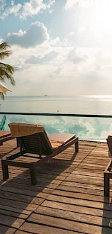 Poolside scene with lounge chairs, umbrellas, palm trees, overlooking an infinity pool blending into a scenic horizon and cloudy sky.