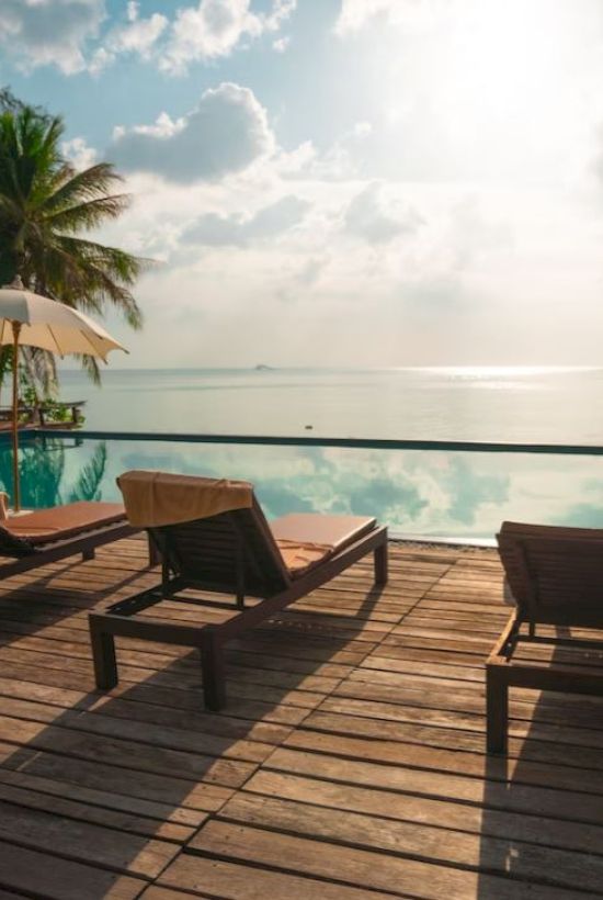 Poolside scene with lounge chairs, umbrellas, and palm trees overlooking a serene body of water under a cloudy sky.