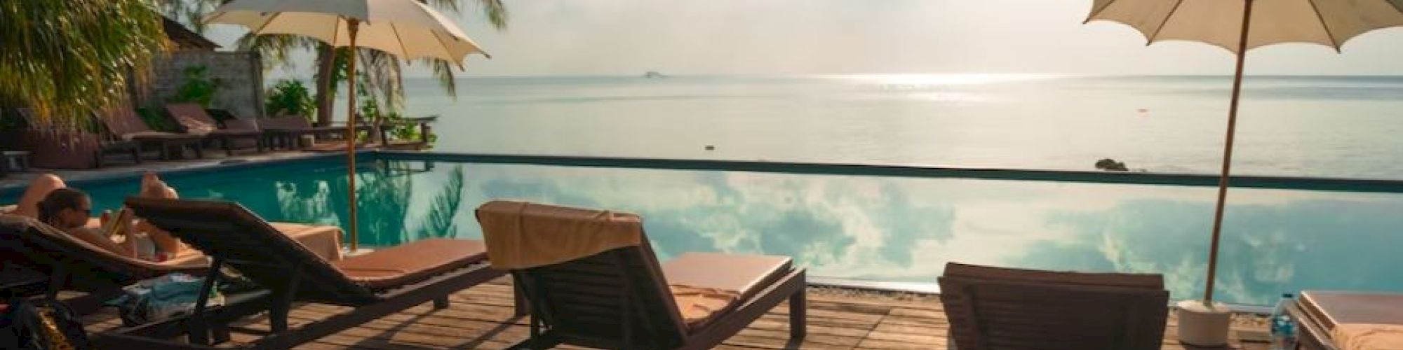 Poolside scene with lounge chairs and umbrellas overlooking a calm ocean, palm trees, and a bright sky with scattered clouds.
