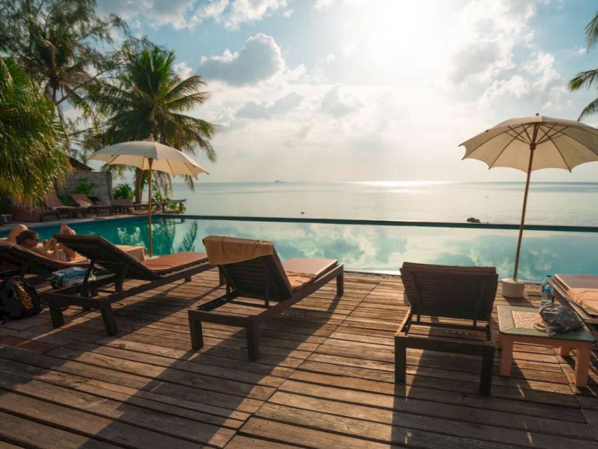 A serene poolside scene with lounge chairs, umbrellas, palm trees, and a view of the sea under a sunny sky, creating a relaxing atmosphere.