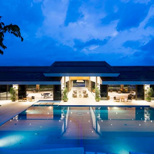 A luxurious, illuminated pool area with a modern building in the background, set against a dusk sky.