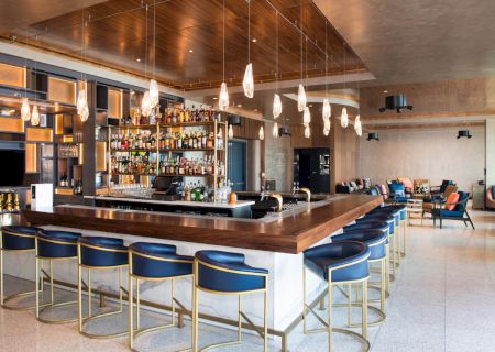 A modern bar with hanging lights, stocked shelves, and blue stools. It features a spacious seating area with couches and a flat-screen TV.