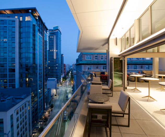 A modern rooftop terrace with tables and chairs overlooks a city street lined with tall buildings, captured during dusk.