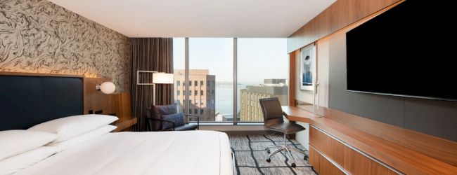 A modern hotel room featuring a large bed, flat-screen TV, desk, and window view of city buildings, with stylish decor and lighting.