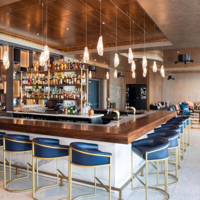 A stylish bar with blue chairs, a wooden counter, hanging lights, and shelves stocked with bottles, set in a spacious, modern lounge.