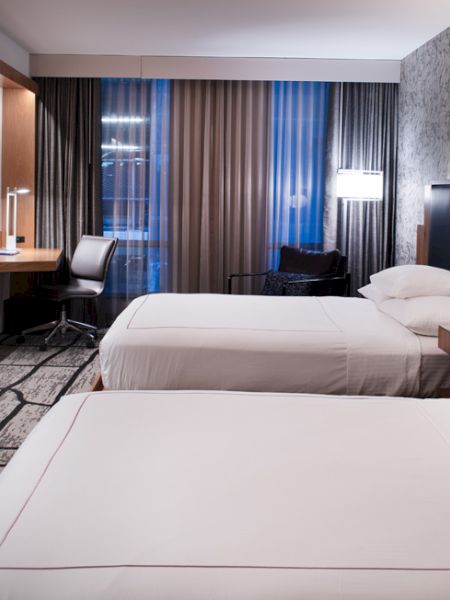 The image shows a modern hotel room with two beds, a TV, a desk, and ambient lighting, featuring a patterned wall and carpet.