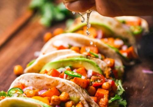 Tacos topped with chickpeas, avocado, and veggies are lined up on a board while lime juice is being squeezed over them.