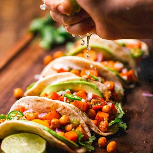 Tacos topped with chickpeas, avocado, and veggies are lined up on a board while lime juice is being squeezed over them.