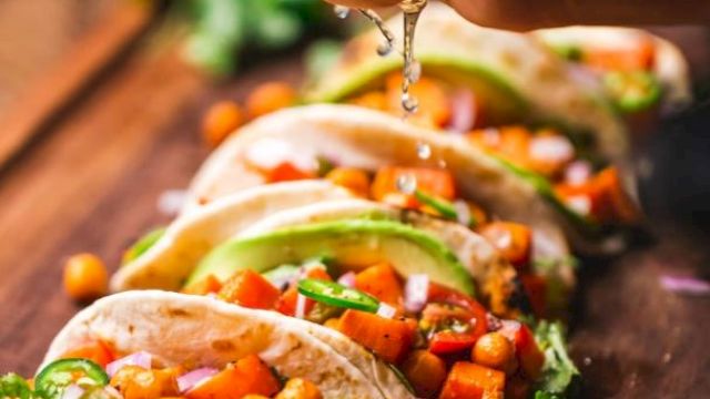 Tacos topped with chickpeas, avocado, and veggies are lined up on a board while lime juice is being squeezed over them.