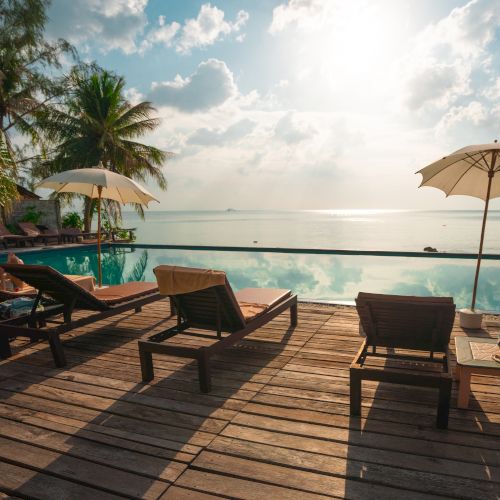 A serene beach scene with loungers and umbrellas on a wooden deck overlooking a calm sea under a partly cloudy sky.