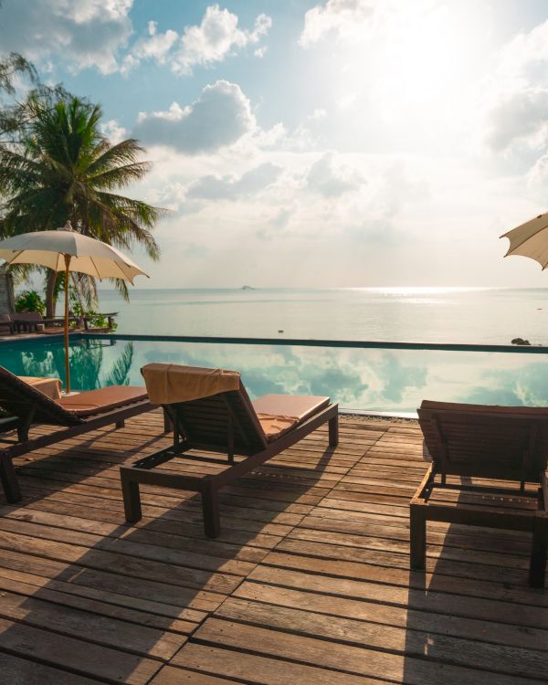 A serene beach scene with loungers and umbrellas on a wooden deck overlooking a calm sea under a partly cloudy sky.