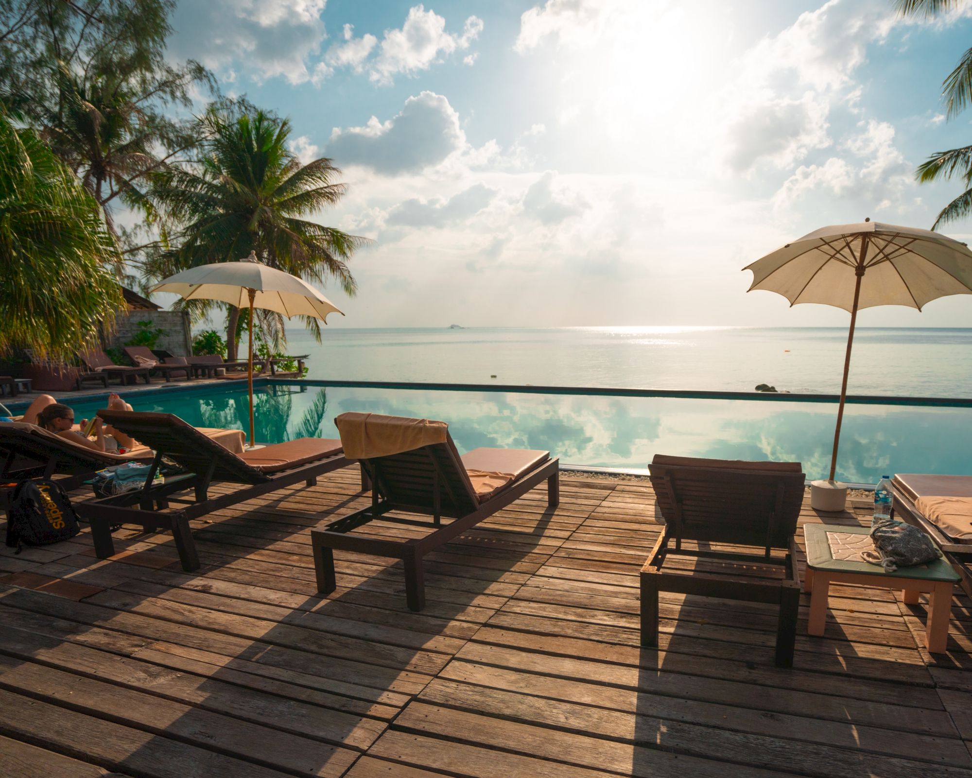 Sun loungers and umbrellas by an infinity pool overlook the ocean, with palm trees and a bright sky in the background.