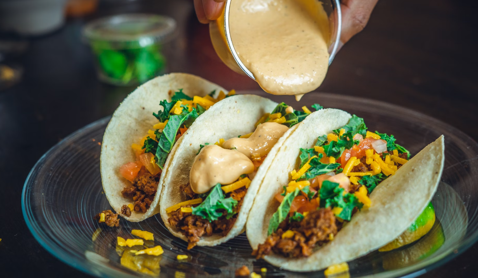 Three tacos with toppings like cheese, lettuce, and sauce being poured from a cup onto the tacos on a plate.