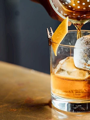 A cocktail being poured over ice in a glass with a lemon twist garnish, on a bar surface.