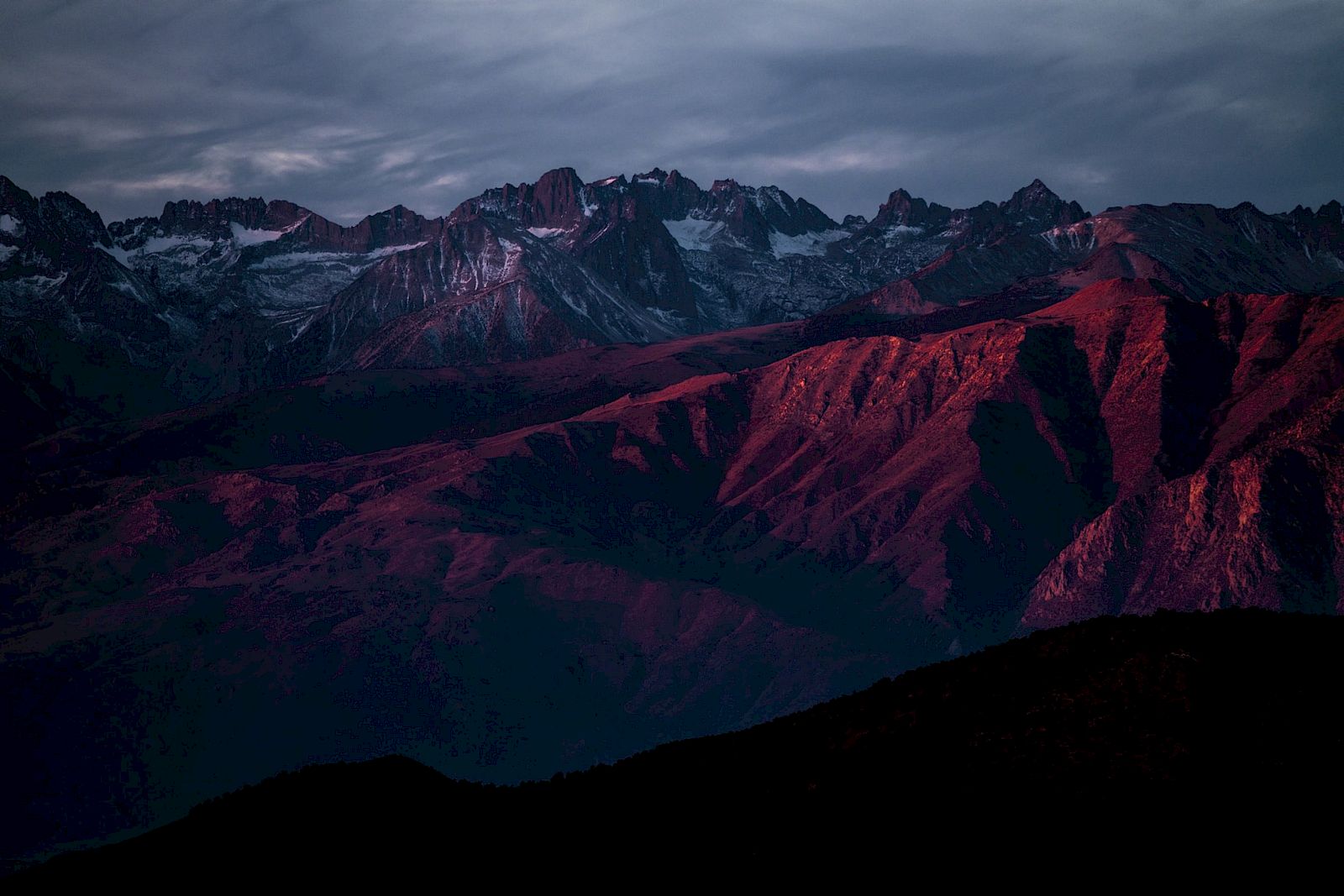 A mountain range with rugged peaks is illuminated by a reddish glow under a cloudy sky.