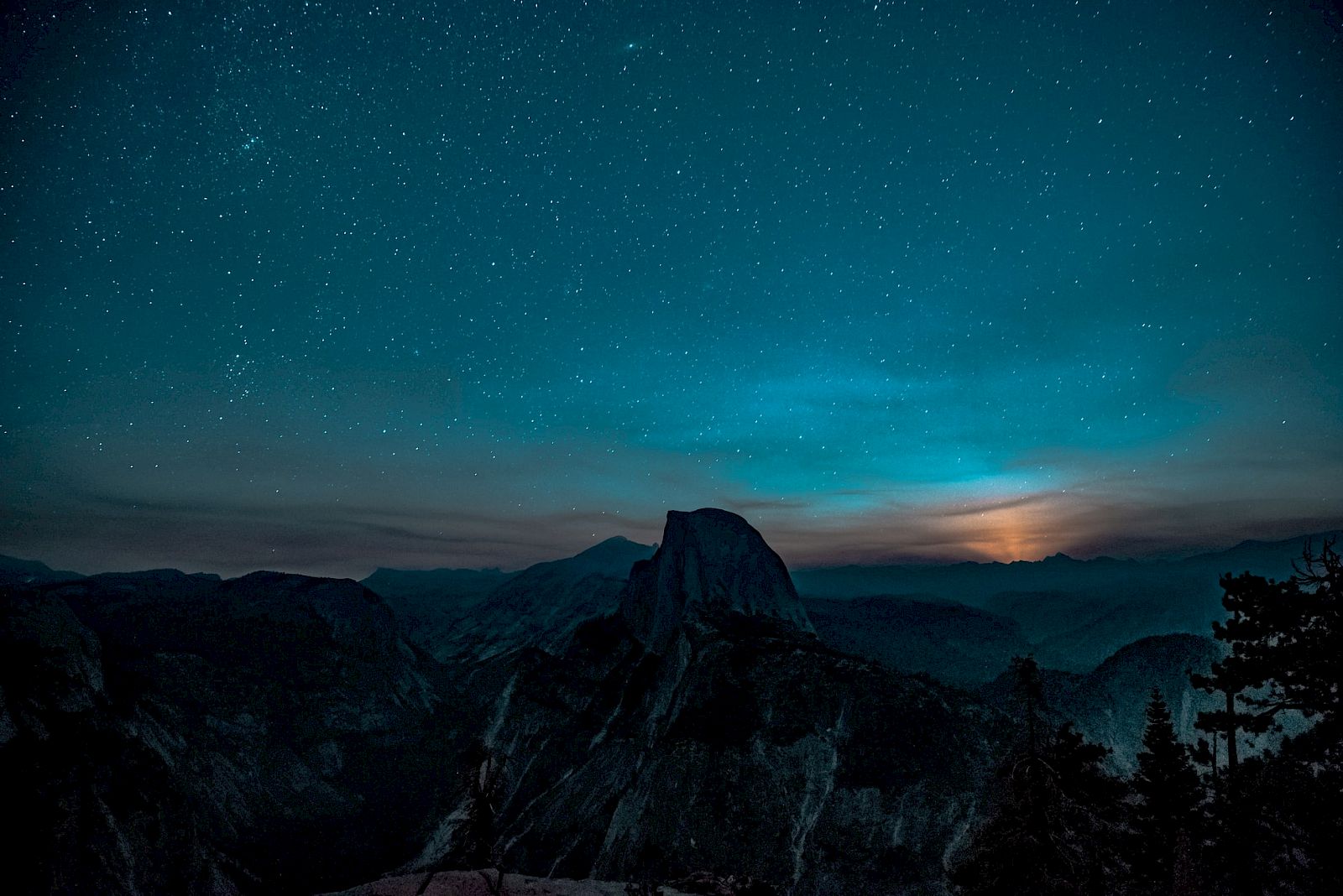 A nighttime landscape shows a starry sky above mountains, with a glow on the horizon and silhouetted trees in the foreground.