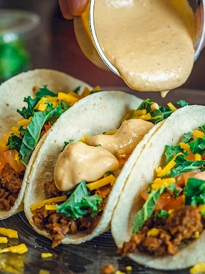 Three tacos with ground meat, cheese, lettuce, tomatoes, and sauce being poured over them, plated on a glass dish.