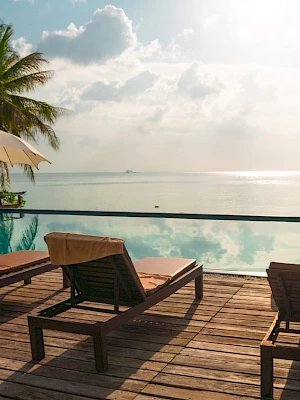 A serene seaside scene with lounge chairs, umbrellas, a swimming pool reflecting clear skies, and palm trees by the ocean at sunset.