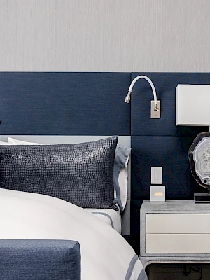 A modern bedroom with a blue and white color scheme, featuring a bed, blue pillows, a lamp on a nightstand, and wall lighting.