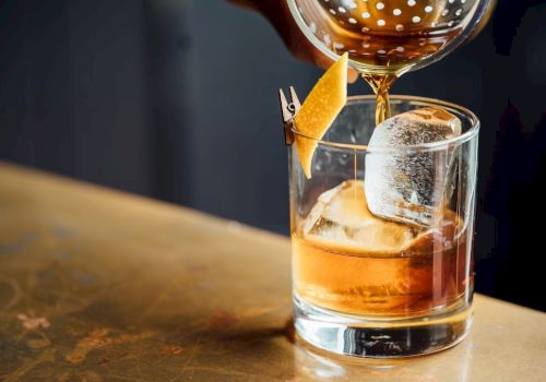A cocktail is being poured into a glass with ice and garnished with a twist of citrus peel.