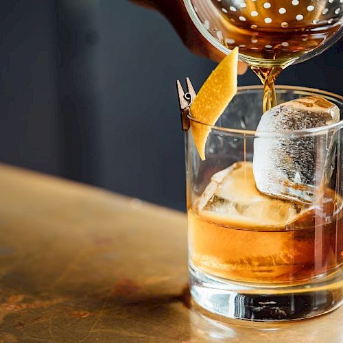A cocktail is being poured into a glass with ice and garnished with a twist of citrus peel.