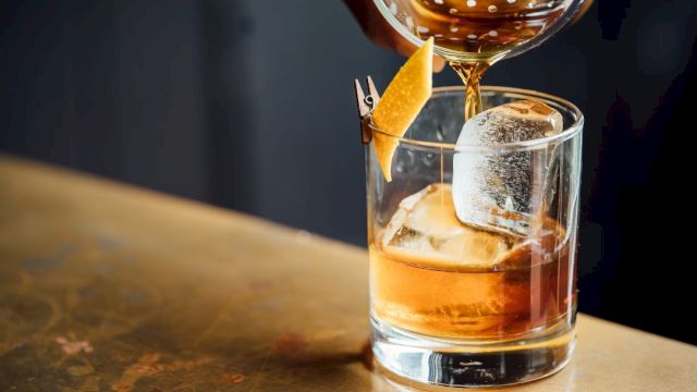 A cocktail is being poured into a glass with ice and garnished with a twist of citrus peel.