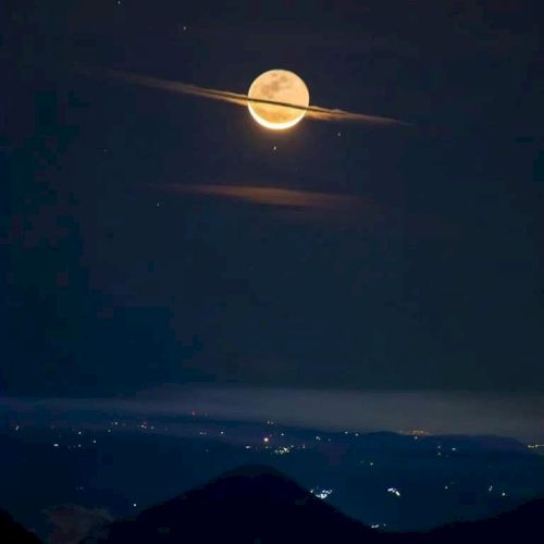 The image shows a full moon with clouds creating a ring-like effect, resembling Saturn, above a landscape with distant city lights.
