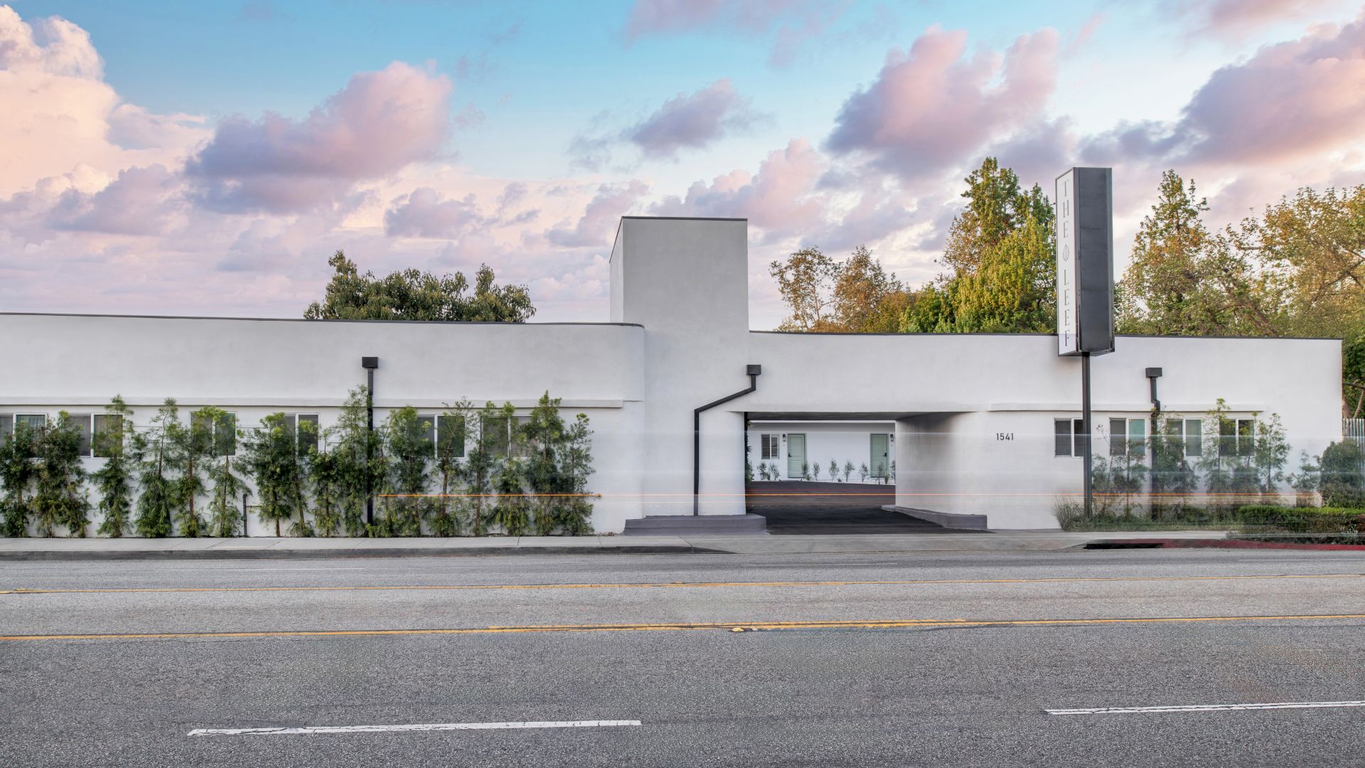 The image shows a modern, white building with greenery and a clear sky backdrop.