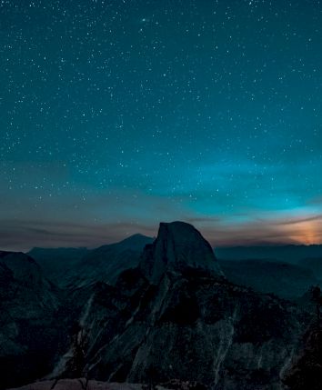 A mountain landscape under a starry night sky, with a glow on the horizon.
