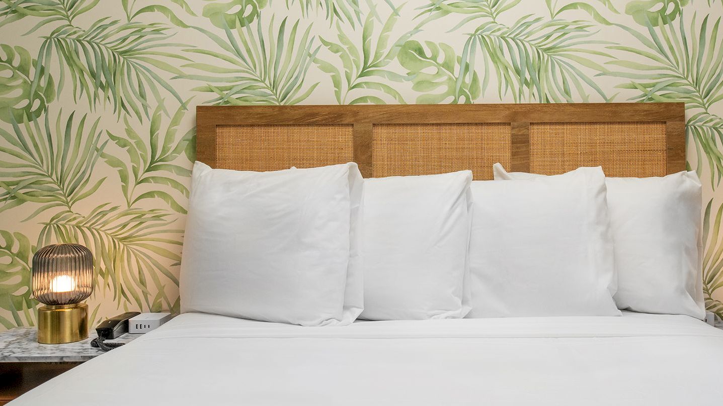 A bed with white pillows, wooden headboard, leafy patterned wallpaper, marble nightstands, and modern lamps.