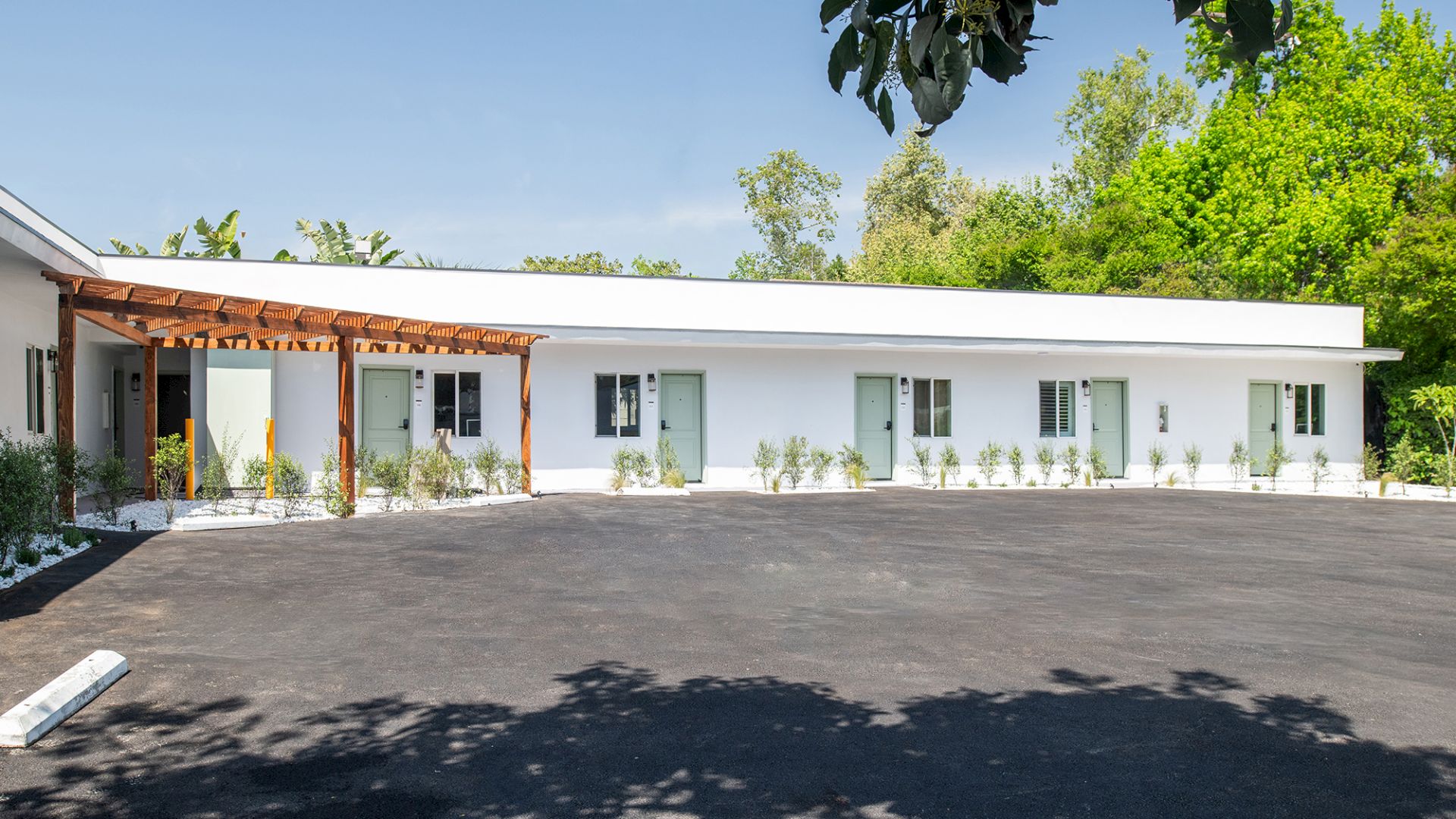 A long, white, single-story building with a wooden pergola and several windows, surrounded by greenery, with a paved area in front.