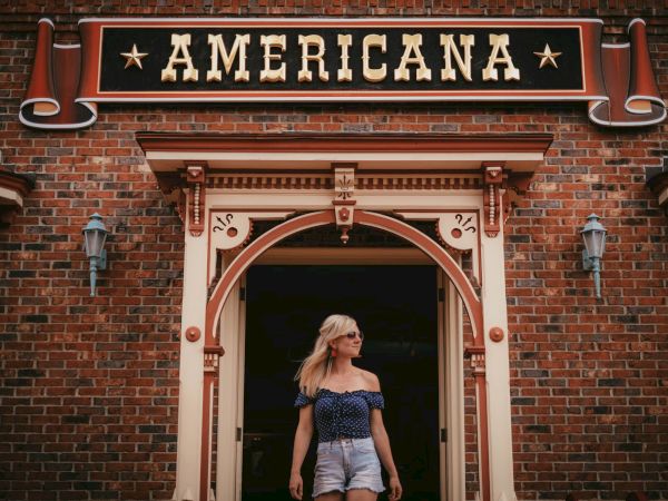 A person is standing under a sign reading "Americana" on a brick building, wearing sunglasses and casual summer clothing.