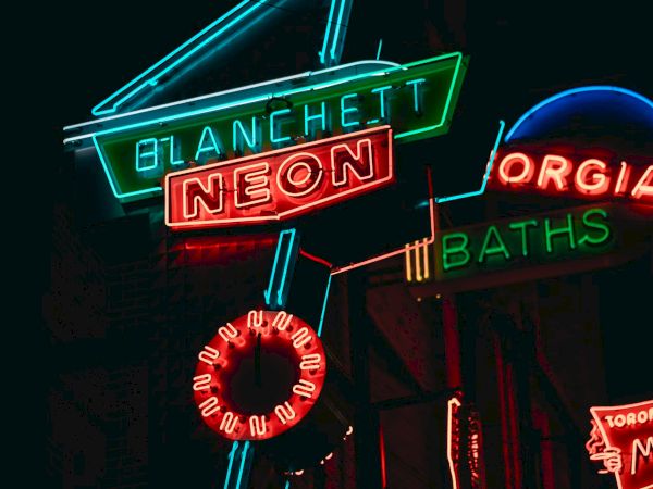 The image shows colorful neon signs with text including "BLANCHETT NEON" and "BATHS," glowing in a dark background.