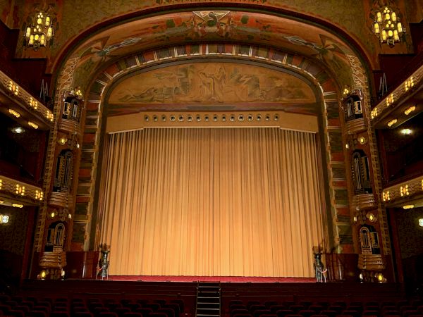 The image shows an elegant, classic theater stage with a closed curtain, surrounded by ornate architectural details and empty seating.