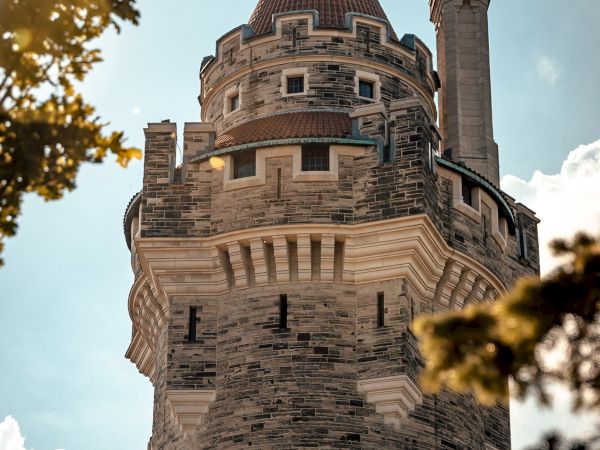 The image shows a stone castle tower with a pointed red roof and decorative windows, framed by green leaves and under a blue sky.