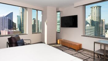 A modern hotel room with large windows showing a cityscape, a bed, TV, and seating area.
