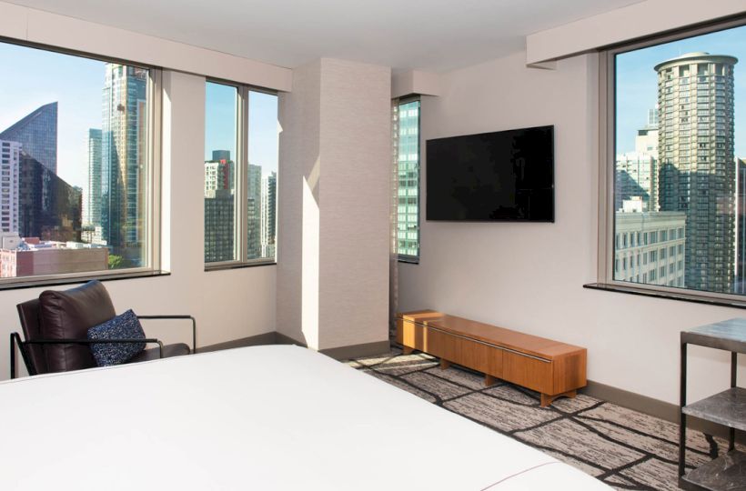 A modern hotel room with large windows showing a cityscape, a bed, TV, and seating area.