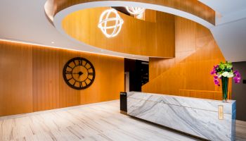 A modern lobby with a marble reception desk, spiral wooden design, large clock on the wall, and a bouquet of flowers on the counter.