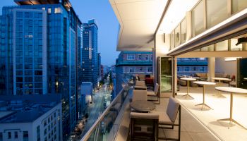 Rooftop terrace at dusk with seating, overlooking a city street, surrounded by tall buildings and illuminated by warm interior lights.