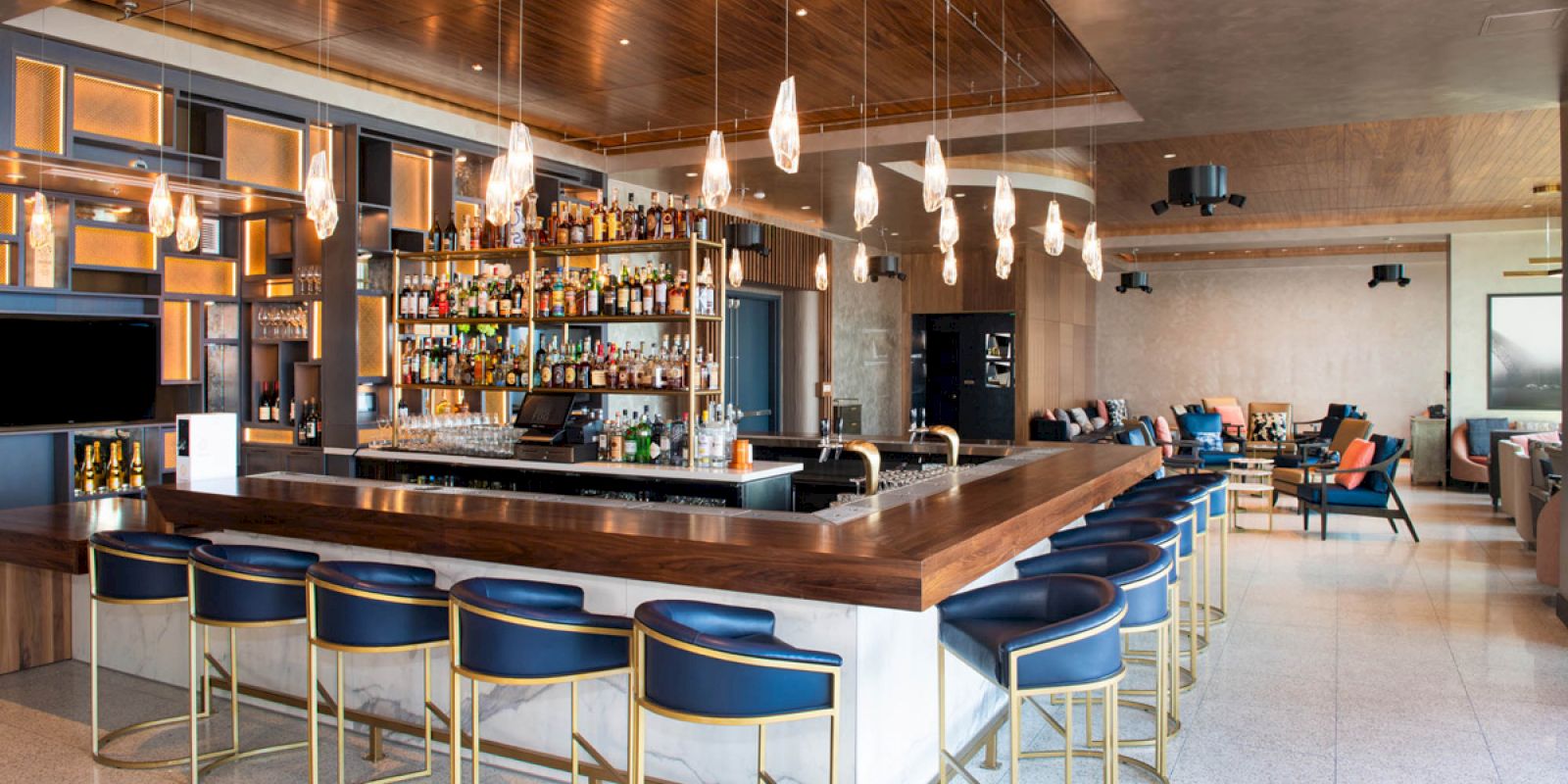 A modern bar features a wooden countertop, blue cushioned stools, pendant lights, and shelves stocked with various bottles in a stylish setting.