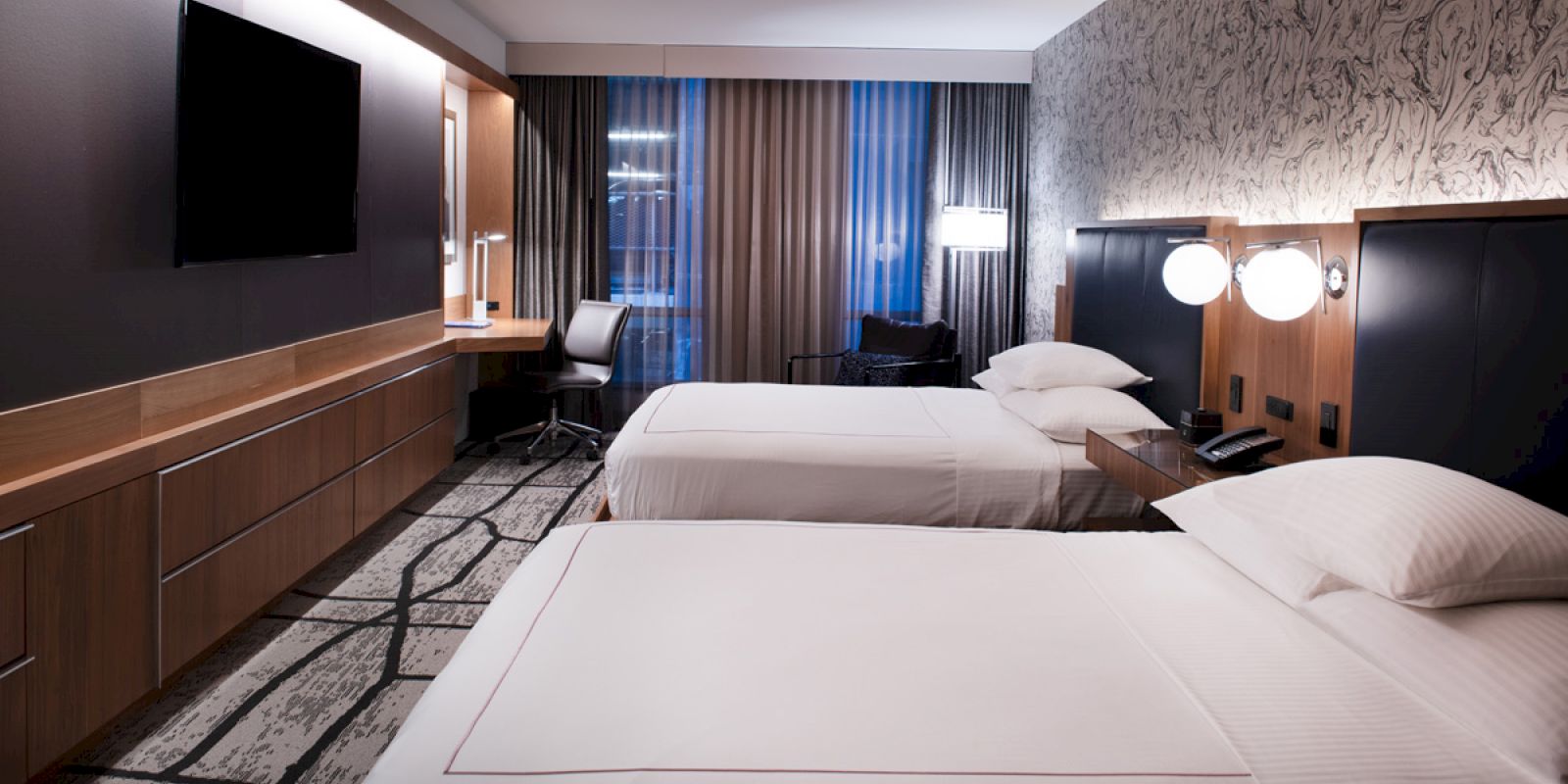 The image shows a modern hotel room with two beds, a TV, a desk, and stylish lighting and decor.