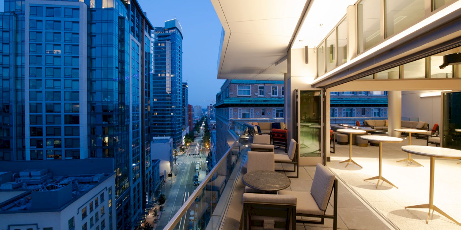The image shows a modern balcony view overlooking a cityscape at dusk, with buildings, outdoor seating, tables, and ambient lighting.
