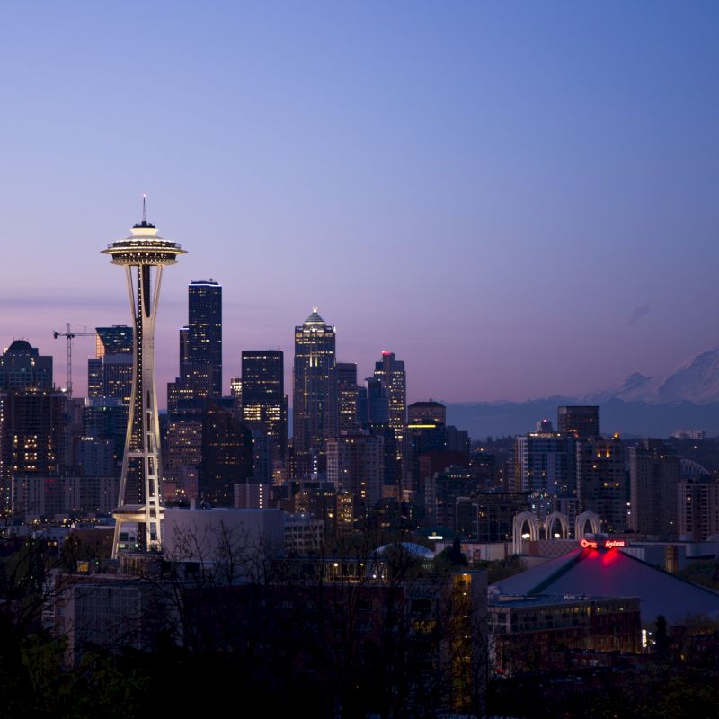 Seattle skyline at dusk featuring the Space Needle, with Mount Rainier in the background against a purple sky.