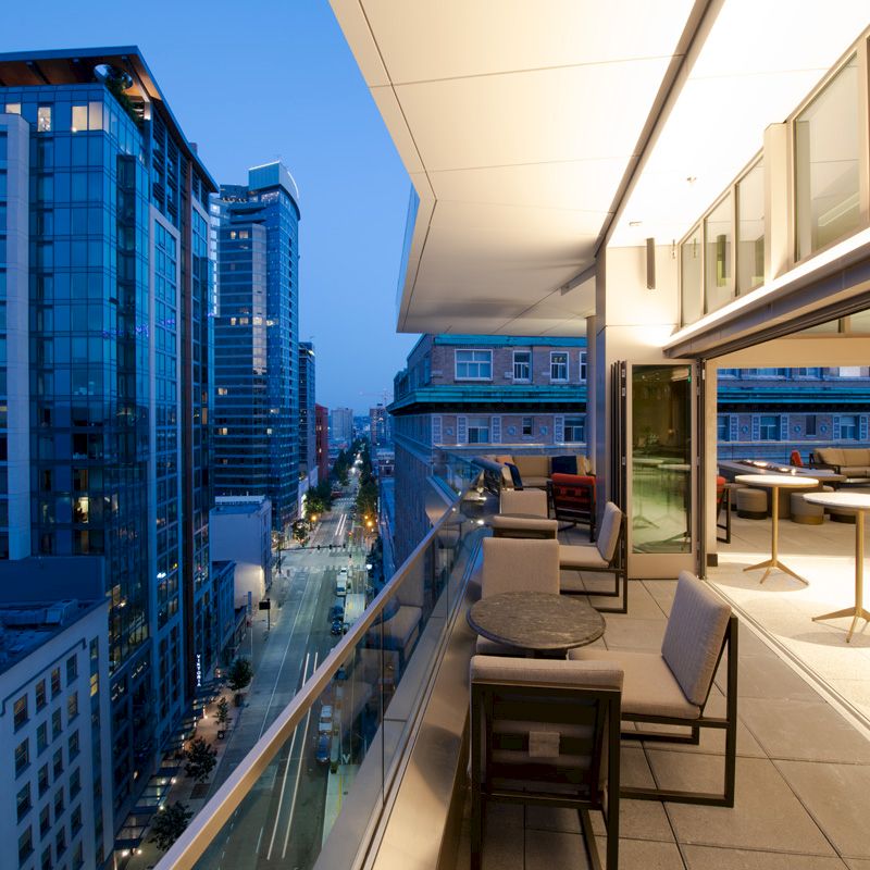 A modern outdoor balcony at dusk, overlooking a city street with high-rise buildings; stylish furniture is arranged for seating.
