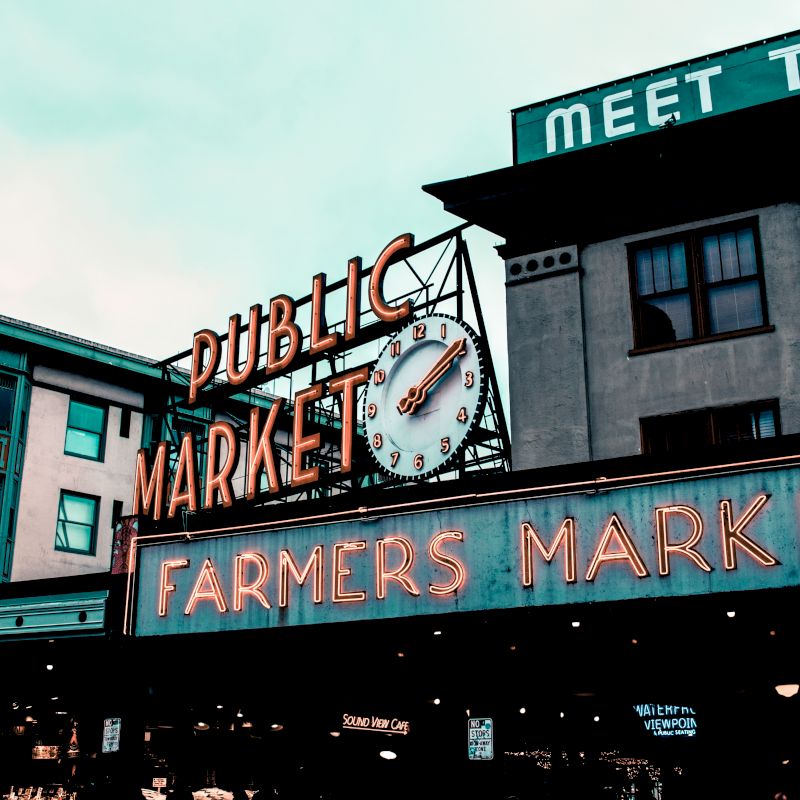 A public market sign with a clock advertising a farmers market, located on a building against a cloudy sky.