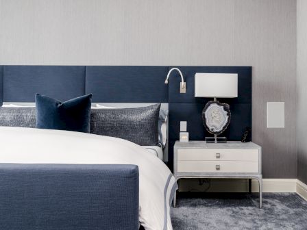 A modern bedroom with a blue upholstered bed, a gray carpet, and a white nightstand featuring a lamp and decorative item.