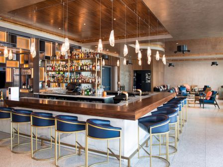A modern bar with a central counter, blue bar stools, hanging lights, and shelves stocked with bottles. Seating area in the background.