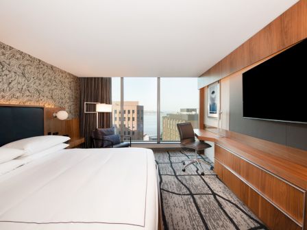 A modern hotel room with a bed, TV, desk, and large window view of buildings and water.